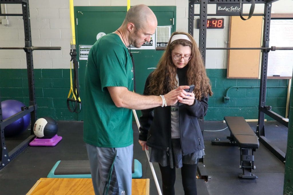 A teacher helps a student with the PLT4M app during a physical education class.