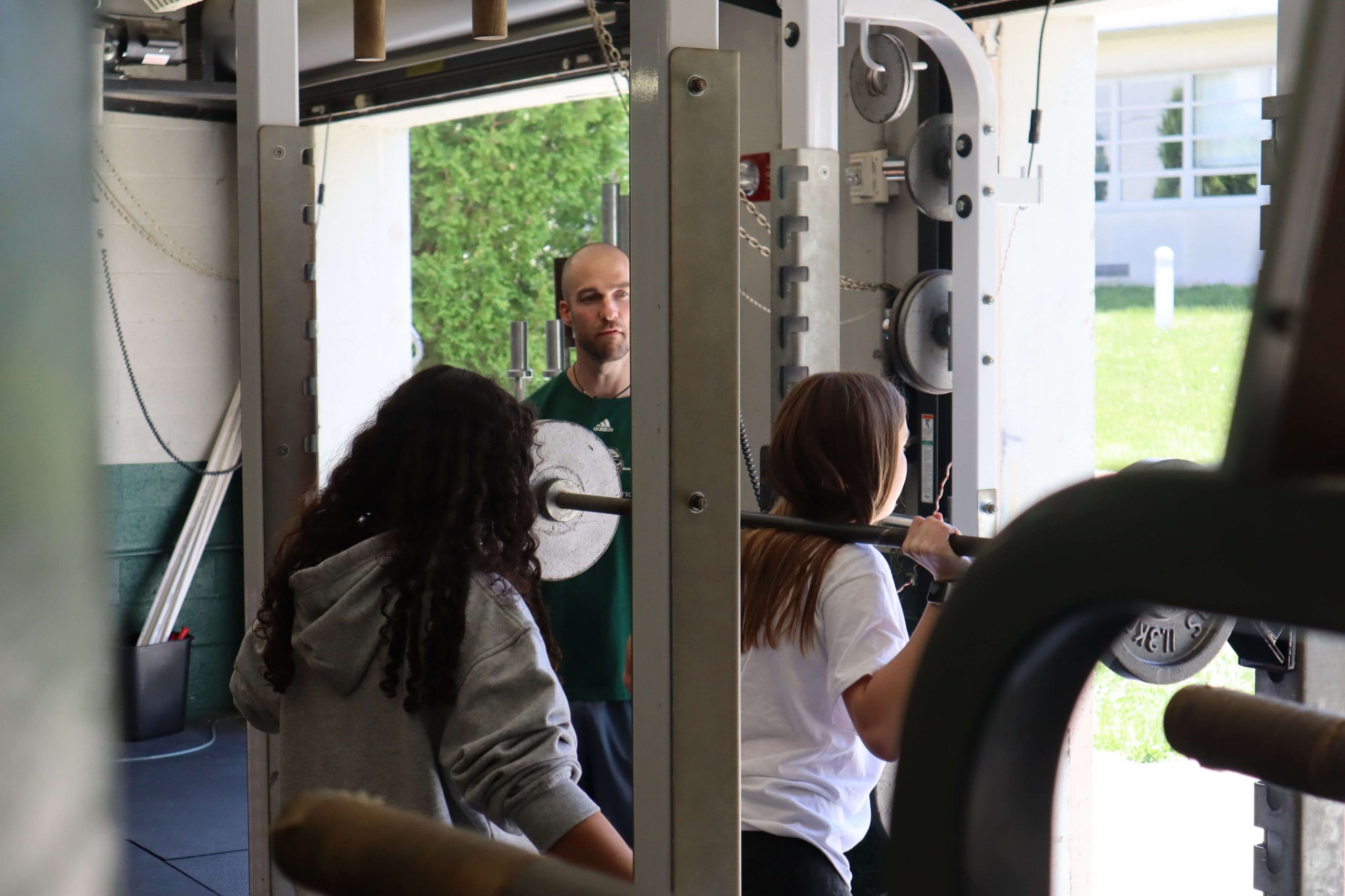 A teacher looks on as a student performs a barbell back squat.