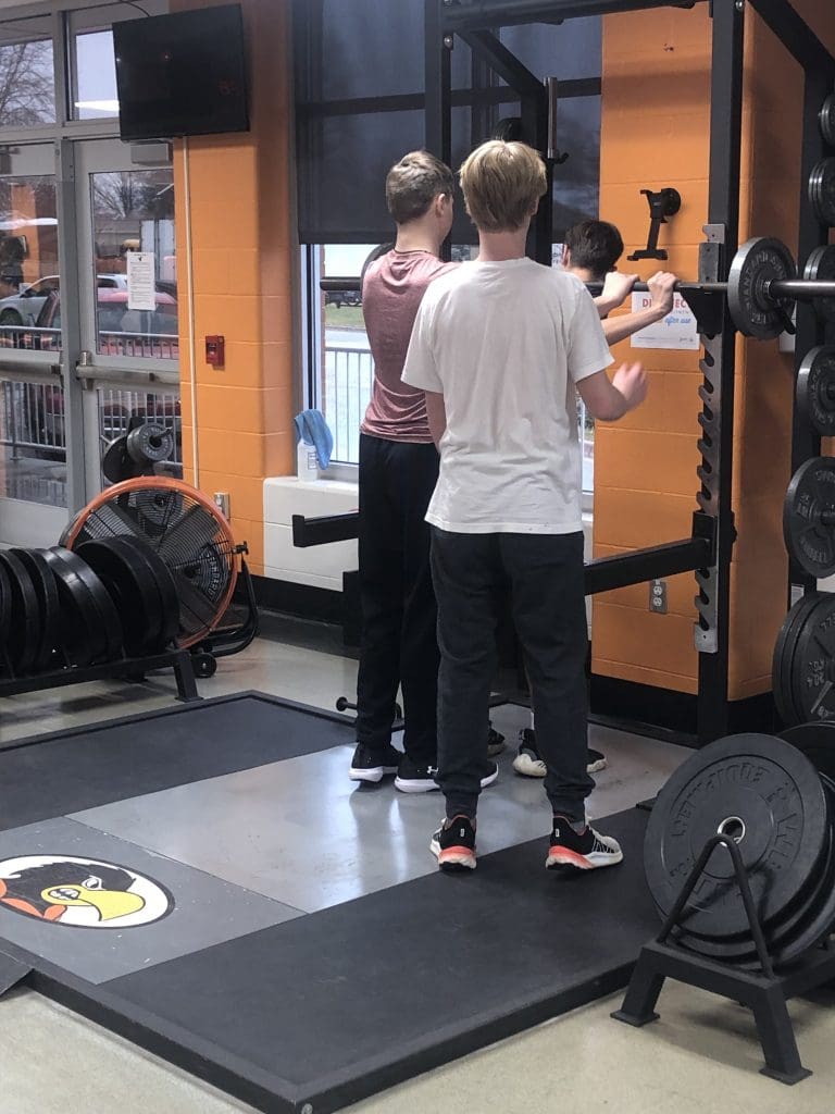 Students help another student safely return a barbell into the weight rack.