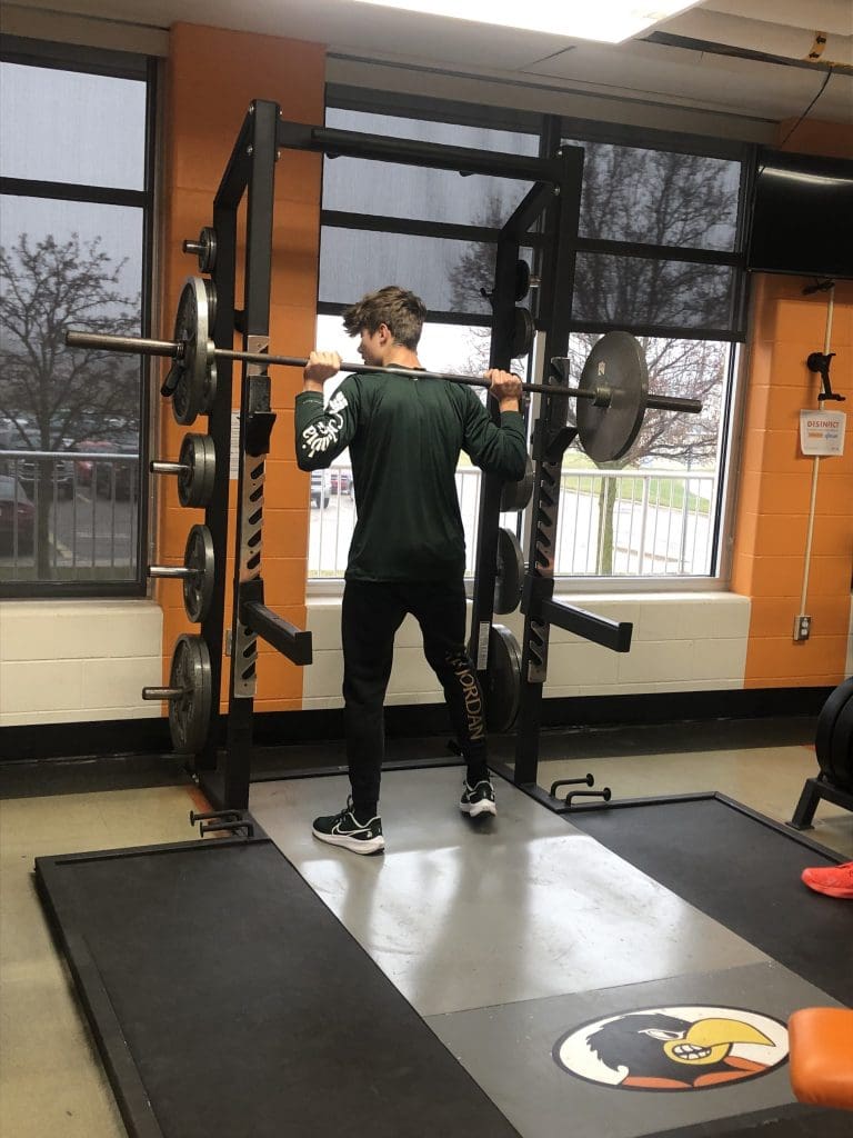 A student steps out of a weight rack with a barbell on his back.