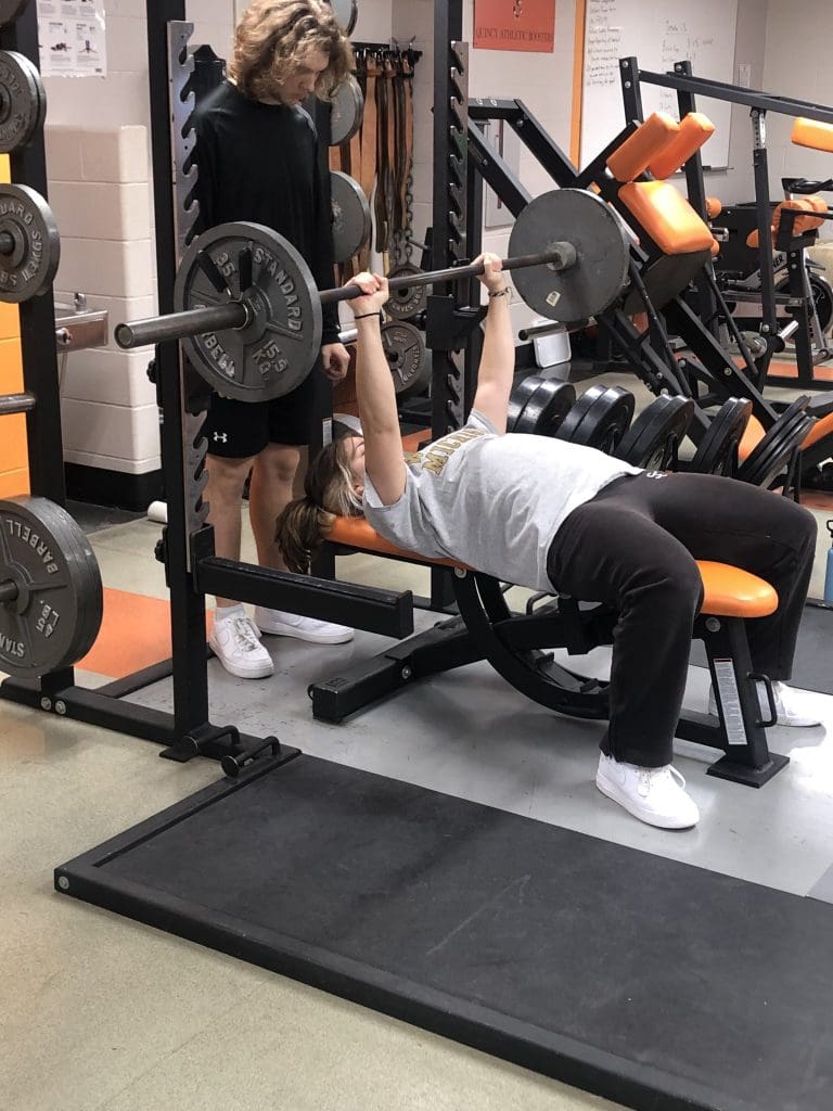 A student performs a bench press exercise while another spots them.