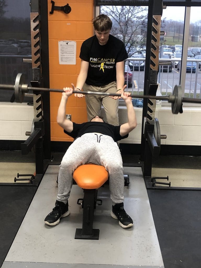 A student performs a bench press rep while another student spots them.