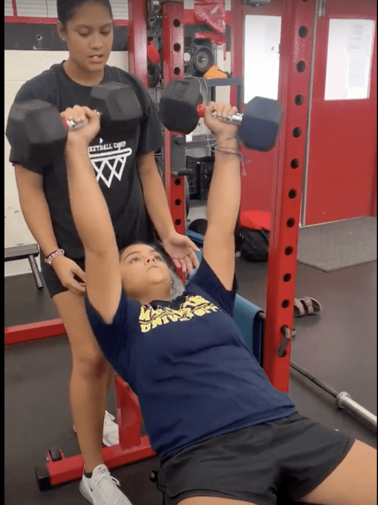 A student performs an incline dumbbell bench press while a classmate spots them during a strength and conditioning physical education class.
