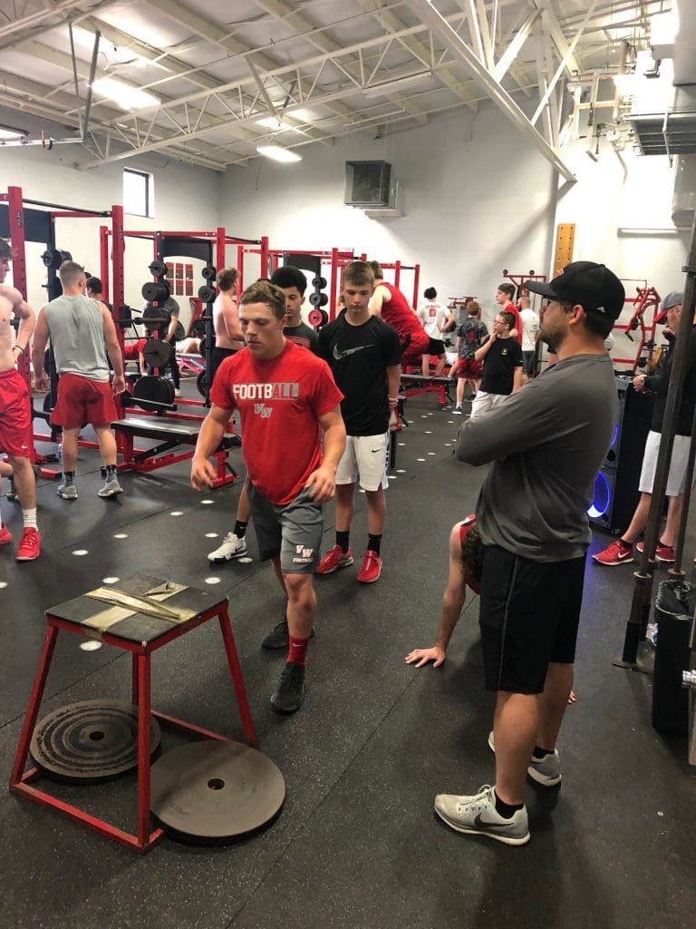A coach looks over a full weight room as an athlete prepares to do a box jump.