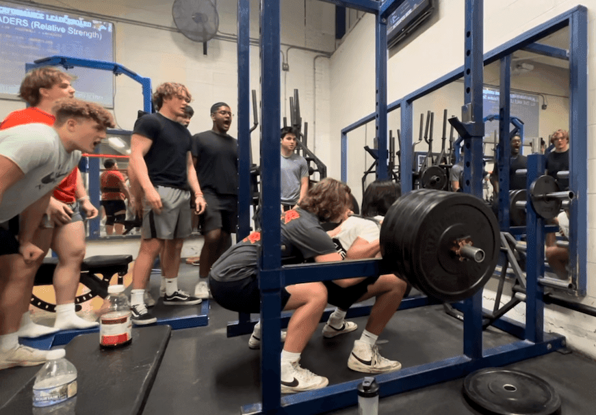 Students cheer on student performing a barbell back squat at naperville north high school in a strength and performance class.